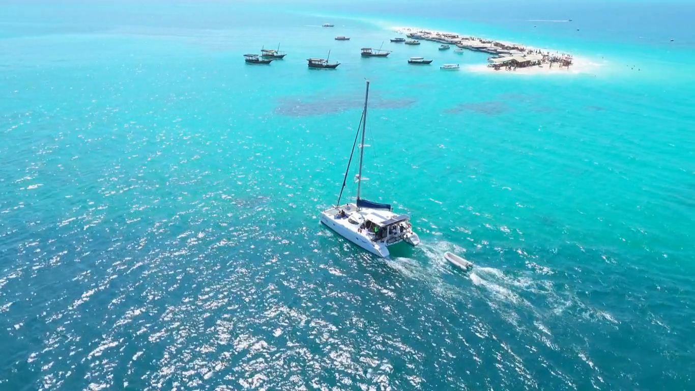 Aerial view of a catamaran crossing turquoise water beside a Zanzibar sandbank